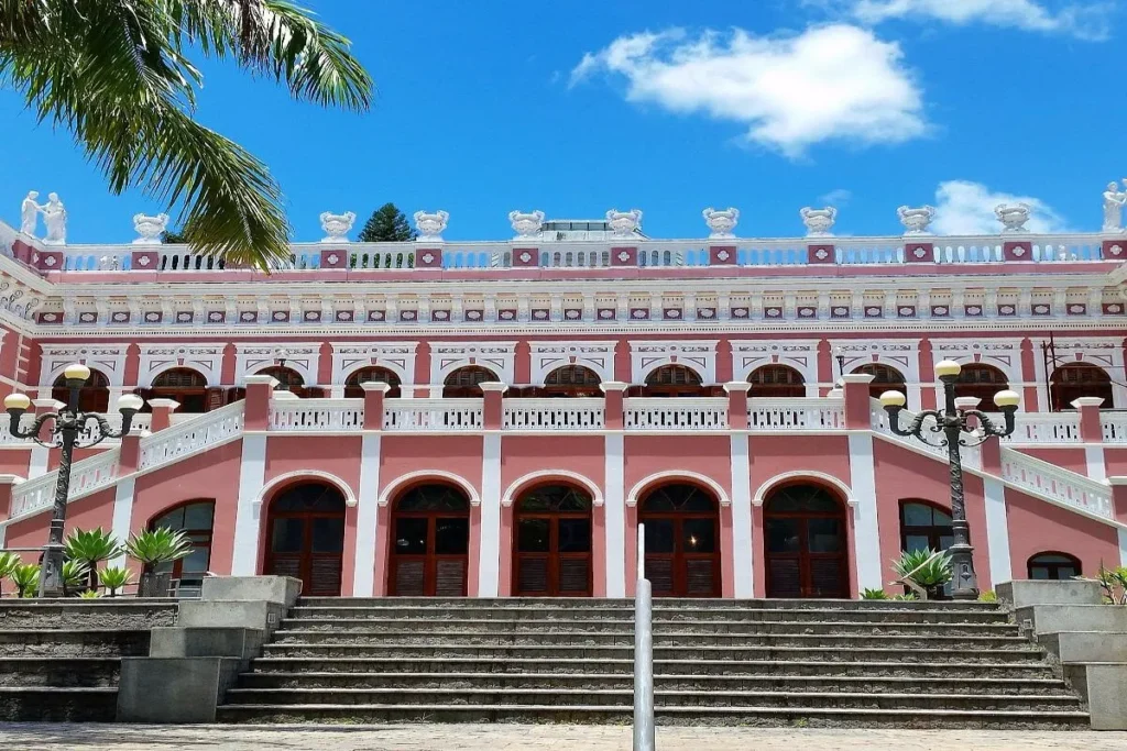 Uma foto frontal do Palácio Rosado em Florianópolis, Brasil. O edifício, de cor rosa e detalhes em branco, possui uma escadaria central que leva a uma varanda no primeiro andar. Vários arcos decoram as janelas e portas. Há dois lampiões antigos em cada lado da escadaria e uma palmeira à esquerda. O céu está azul com algumas nuvens brancas, e o sol ilumina a fachada do prédio. A arquitetura é um exemplo do estilo neoclássico.