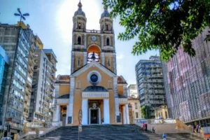 Uma foto da Catedral Metropolitana de Florianópolis, no Brasil. O edifício de cor amarela e branca, em estilo neoclássico, domina o centro da imagem. Há duas torres sineiras e uma grande escadaria na frente. A foto foi tirada em um dia de sol, com o brilho do sol no alto da imagem. A catedral é cercada por prédios modernos e altos.
