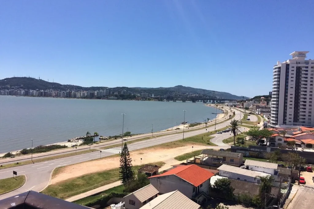 Uma foto aérea da orla de Florianópolis, Brasil, com a Baía Sul e a Ponte Hercílio Luz ao fundo. Uma rodovia costeira segue a curva da baía, e no primeiro plano, há prédios residenciais. Montanhas cobertas de vegetação formam o horizonte. O céu está azul e ensolarado.