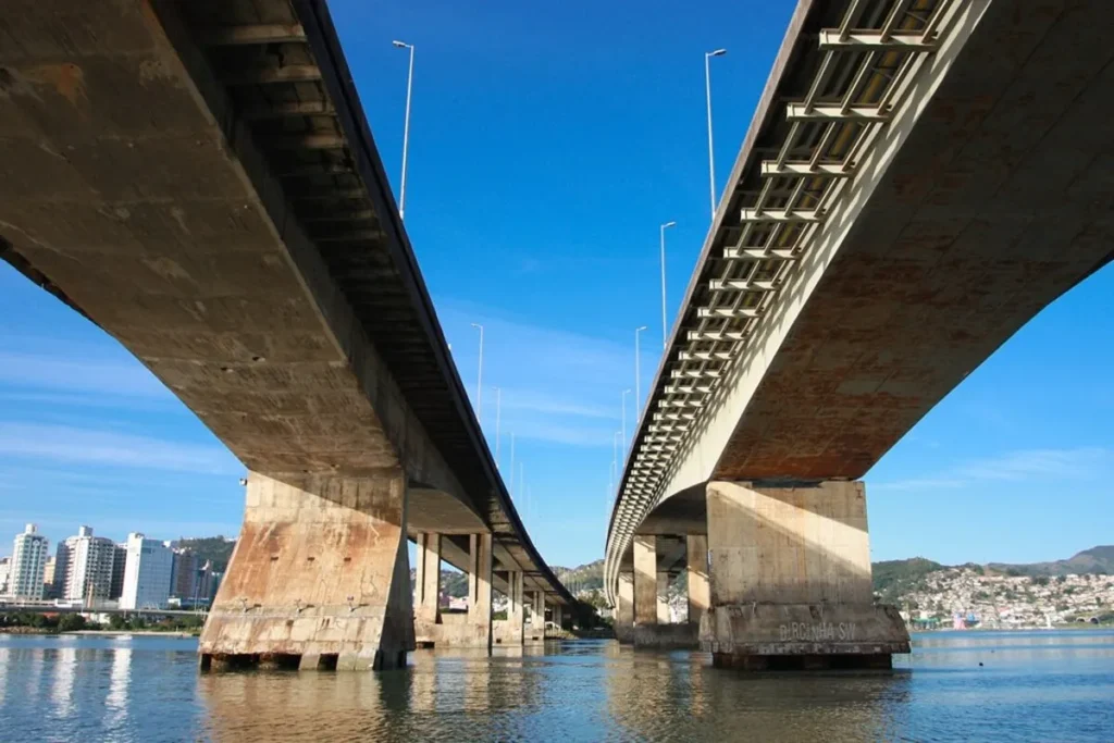 Uma foto da Ponte Hercílio Luz em Florianópolis, Brasil, vista do nível da água. As duas grandes torres de ferro cinza da ponte e o sistema de cabos suspensos se destacam. O céu está azul e a água da baía reflete a estrutura da ponte. A imagem transmite a imponência da engenharia histórica.