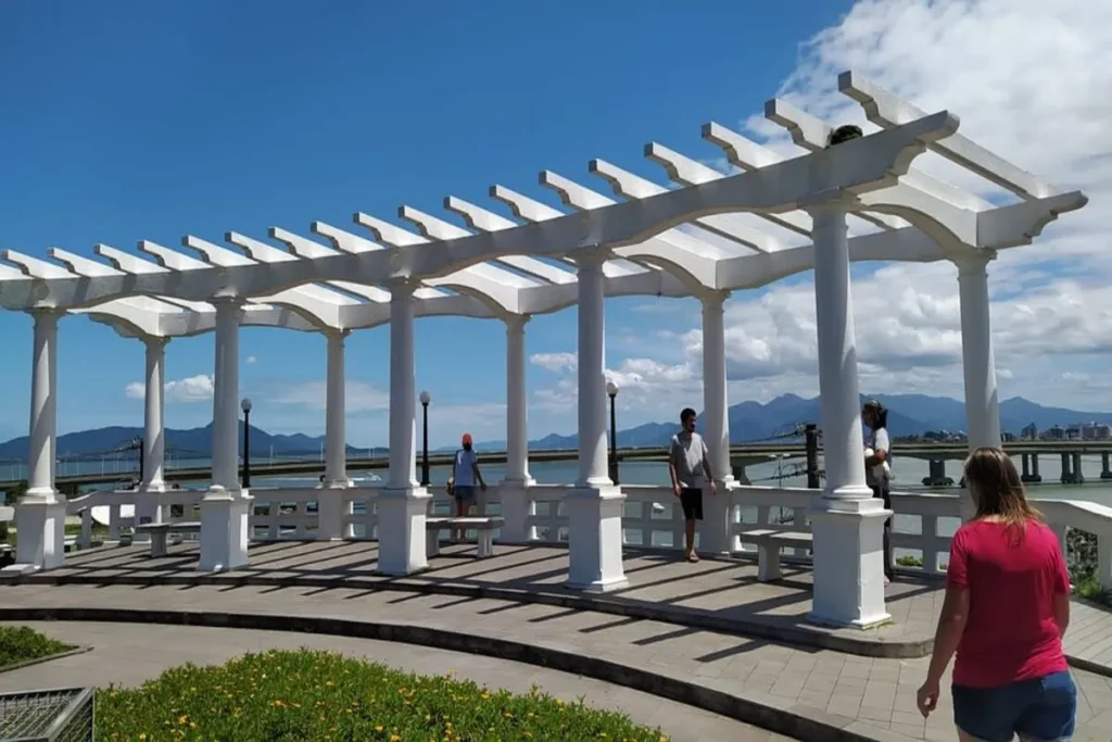 Pergolado branco no Parque da Luz, em Florianópolis, com vista para o mar e montanhas.