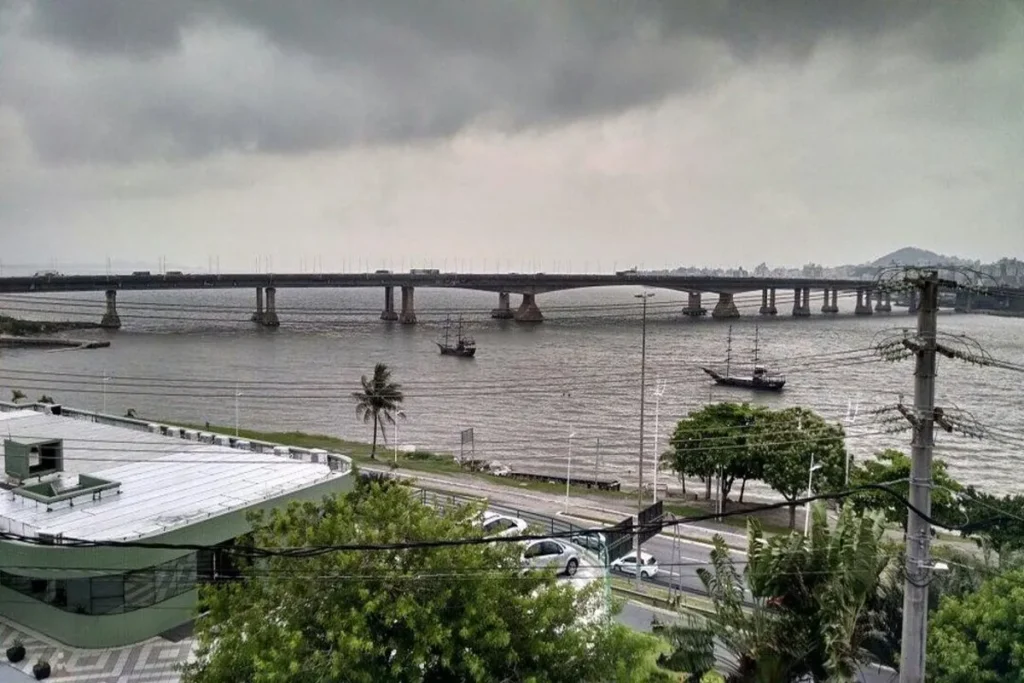 Ponte Hercílio Luz em Florianópolis sob céu nublado, com dois barcos no mar e prédios na costa.