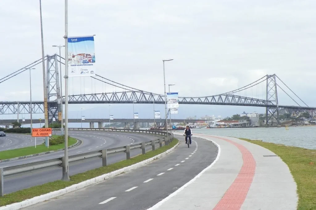 Vista aérea da Ponte Hercílio Luz em Florianópolis, Brasil, com ciclovia e rodovia em primeiro plano, e a baía ao lado. Um ciclista pedala pela ciclovia, e edifícios podem ser vistos à distância.