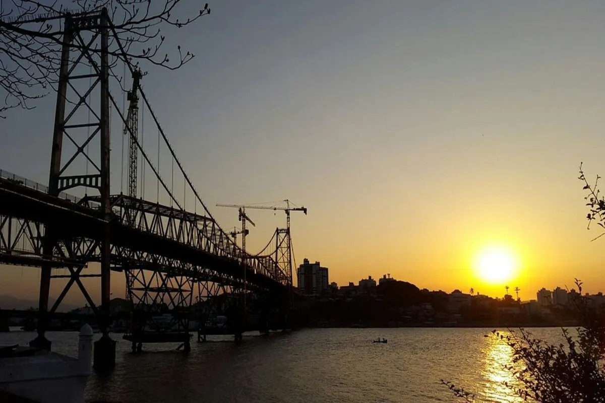 Silhueta da Ponte Hercílio Luz ao pôr do sol, com o sol laranja se pondo sobre a cidade.