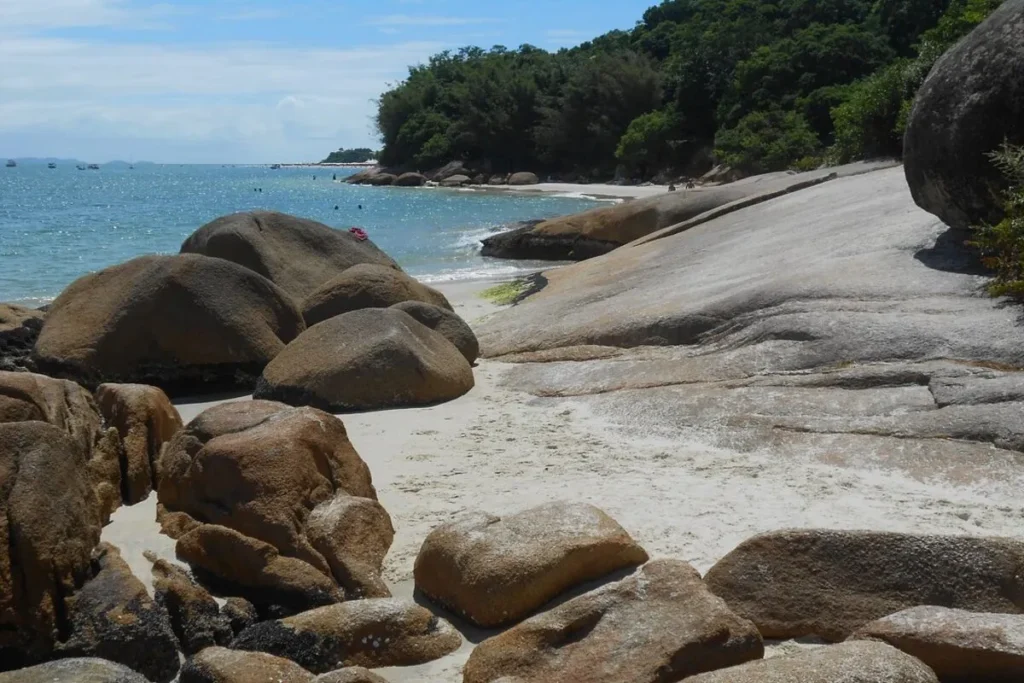 Formações rochosas na areia da Praia da Daniela com mar azul e vegetação ao fundo.