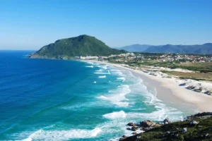 Vista aérea da Praia do Santinho em Florianópolis com mar azul, dunas e morro verde.
