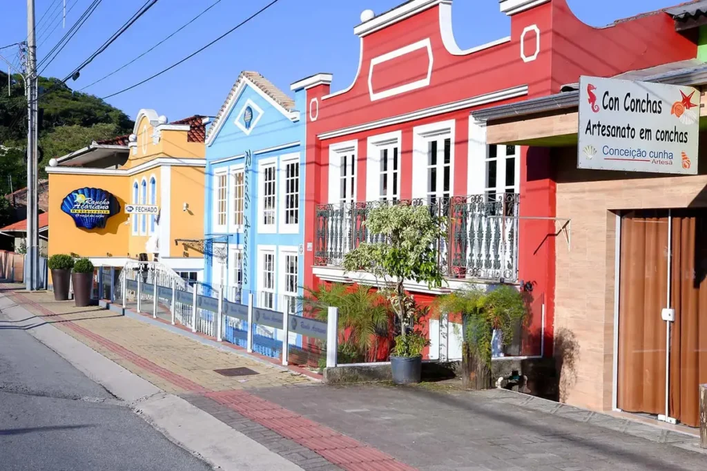 Fileira de casas coloniais coloridas (vermelho, azul, amarelo) e preservadas na rua de Ribeirão da Ilha, com balcões de ferro e placas de artesanato e restaurante.