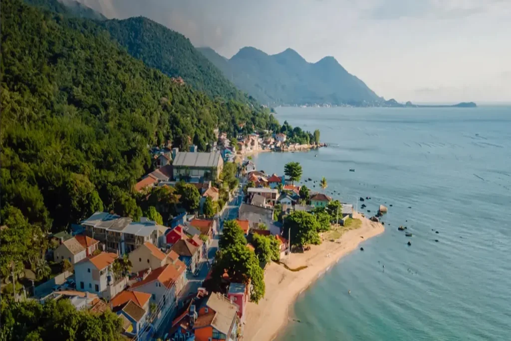 Drone view panorâmica da vila histórica de Ribeirão da Ilha, Florianópolis, mostrando a orla, casario colonial, praia de areia e montanhas cobertas de mata atlântica no fundo.