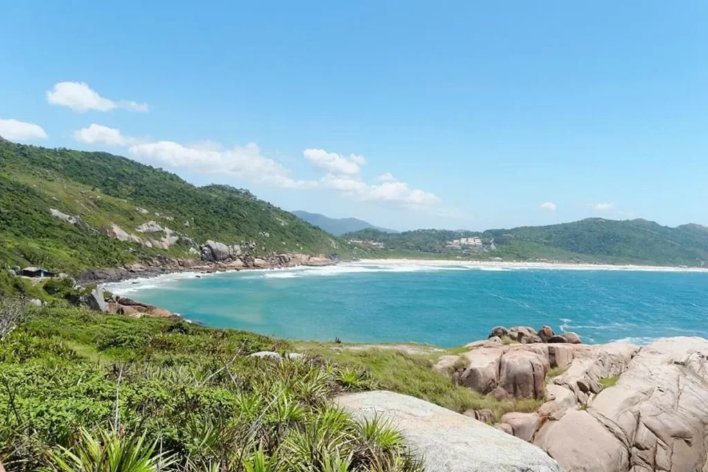 Vista panorâmica de uma baía de águas azul-turquesa, cercada por montanhas verdes e rochas. A imagem mostra a Trilha do Gravatá e, ao fundo, a Praia da Galheta, com uma longa faixa de areia. O céu é azul com nuvens brancas e o sol ilumina a paisagem.