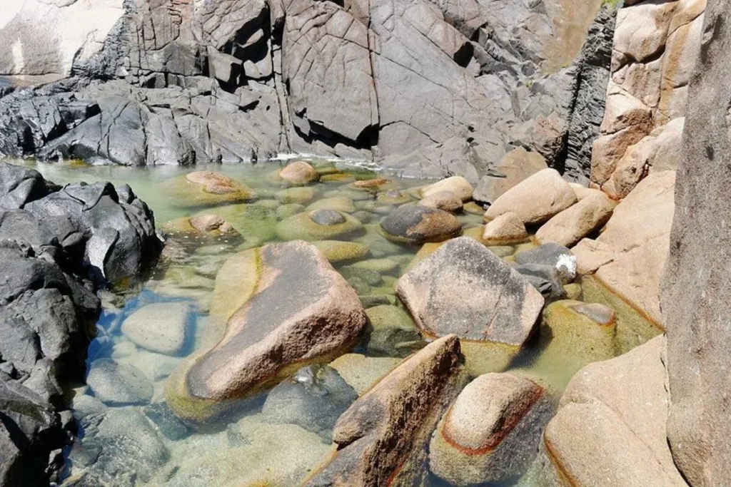 Uma piscina natural de águas claras e rasas, cercada por grandes rochas escuras e cinzas. A água é tão límpida que as pedras no fundo são perfeitamente visíveis. O sol ilumina a paisagem e destaca as diferentes texturas e cores das rochas.