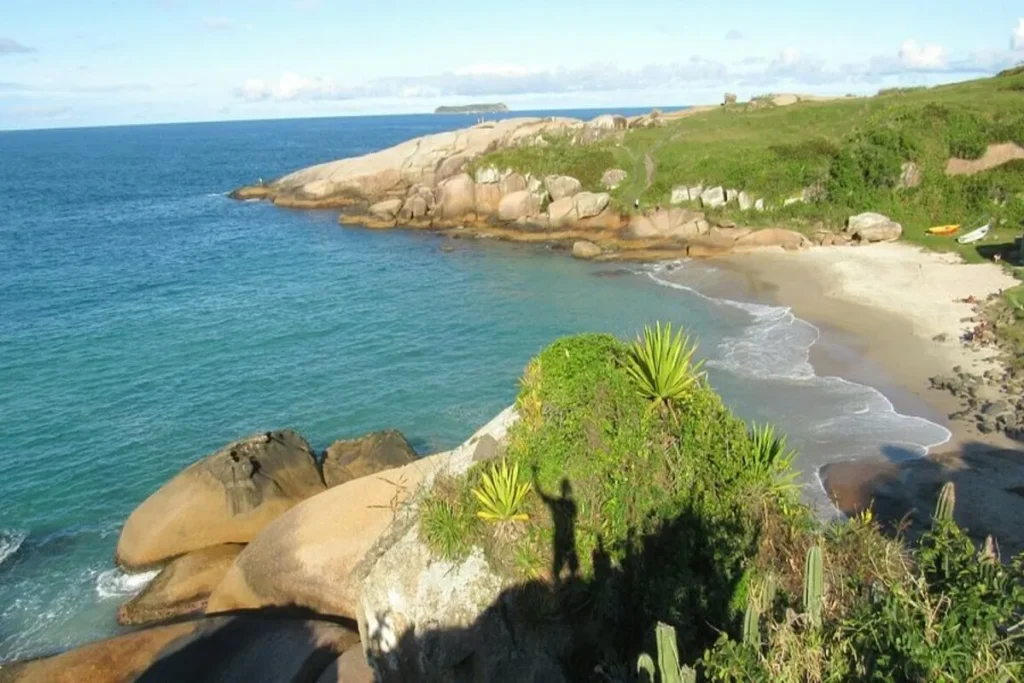 Uma vista panorâmica da costa de Laguna, com a Trilha do Gravatá em primeiro plano e a Praia do Rosa ao longe. O céu é azul com poucas nuvens e a água do oceano é de um tom azul-claro, com ondas suaves que chegam à areia. A paisagem é dominada por colinas gramadas com vegetação nativa e grandes rochas. No horizonte, a Ilha do Batuta e a Ilha do Corvo são visíveis.