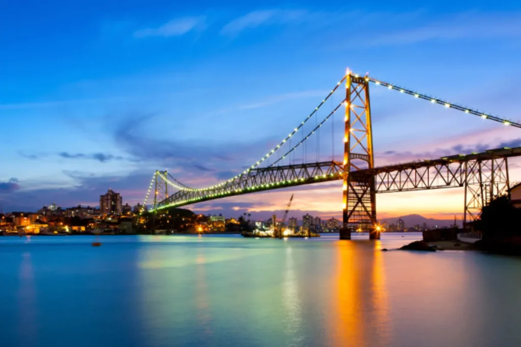 Ponte Hercílio Luz, símbolo de Florianópolis, iluminada durante o pôr do sol com a paisagem urbana ao fundo e o reflexo na água. Ideal para conteúdos sobre a capital catarinense, turismo, engenharia, e pontos turísticos.