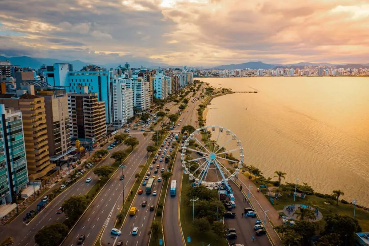 Vista aérea da orla de Florianópolis ao pôr do sol, com uma avenida, prédios, e uma roda-gigante à beira-mar. Ideal para imagens de viagem, paisagens urbanas e turismo em Santa Catarina.