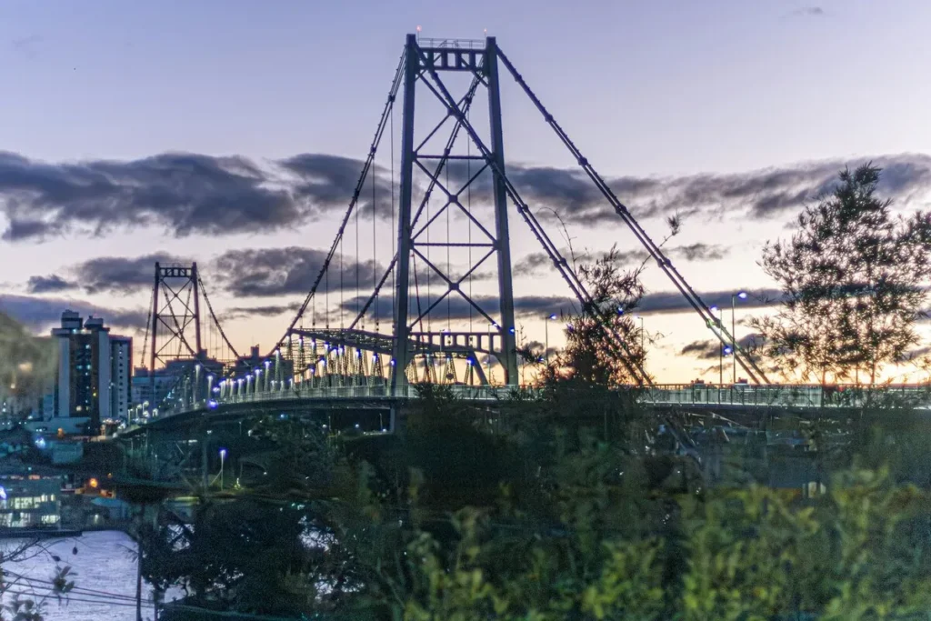 Vista noturna da Ponte Hercílio Luz (estrutura suspensa de aço) em Florianópolis, com luzes acesas, edifícios da cidade no fundo e céu crepuscular, representando a capital e sua infraestrutura.