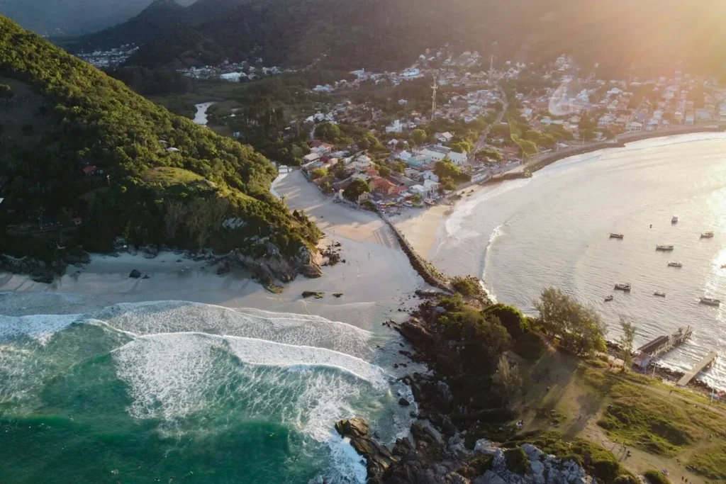 Vista de drone de praia e bairro residencial de Florianópolis ao pôr do sol, mostrando a orla, mar de cor turquesa, montanha e casas próximas ao oceano, que afetam o custo de vida.