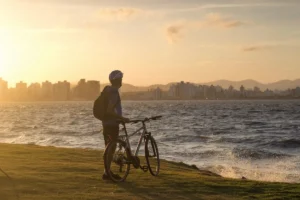 Homem de capacete e mochila com bicicleta, em área gramada na orla da baía de Florianópolis, observando o skyline da cidade e o mar sob a luz dourada do pôr do sol, representando lazer e custo de vida.