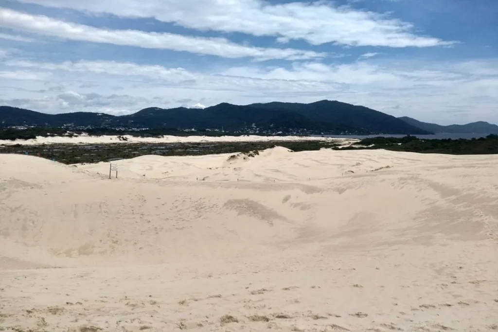 Uma foto das Dunas da Joaquina em Florianópolis, Brasil, com a areia branca e ondulada no primeiro plano. Ao fundo, uma área com vegetação escura leva à Lagoa da Conceição e a uma cadeia de montanhas. O céu está parcialmente nublado com nuvens.