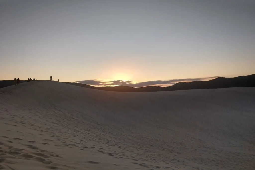 Uma foto das Dunas da Joaquina em Florianópolis, Brasil, ao pôr do sol. A luz dourada do sol se esconde atrás de um morro escuro no horizonte. No primeiro plano, a areia clara das dunas é marcada por pegadas. Algumas silhuetas de pessoas se destacam na crista de uma duna.