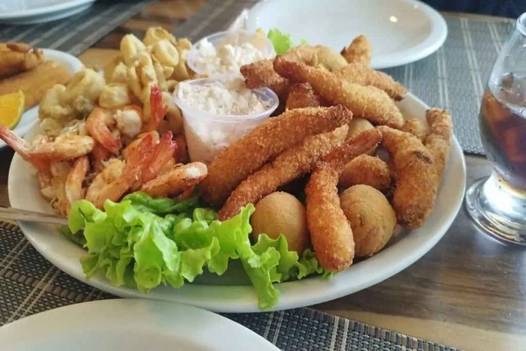 Prato branco com uma variedade de frutos do mar fritos, incluindo camarão e lula, servido com alface e dois potes de molho tártaro. Cena de petiscos de frutos do mar.