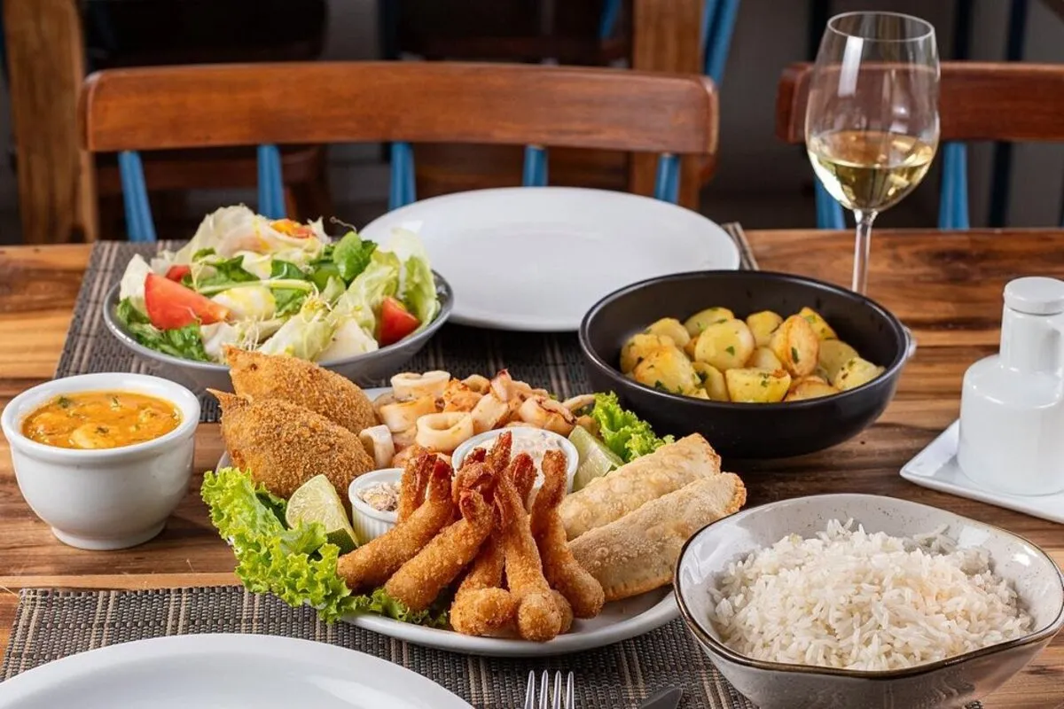 Travessa de frutos do mar fritos (camarão, peixe e lula) servida em uma mesa com acompanhamentos: arroz, batatas, salada e molho. Cena de restaurante de frutos do mar.