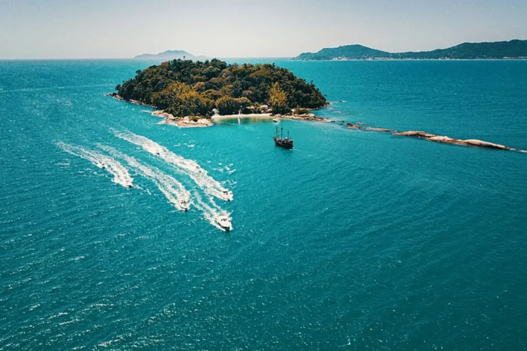 Vista de drone de ilha deserta e coberta de mata atlântica em Florianópolis, com mar de cor turquesa, barco pirata ancorado e lanchas de passeio em alta velocidade deixando rastros de espuma.