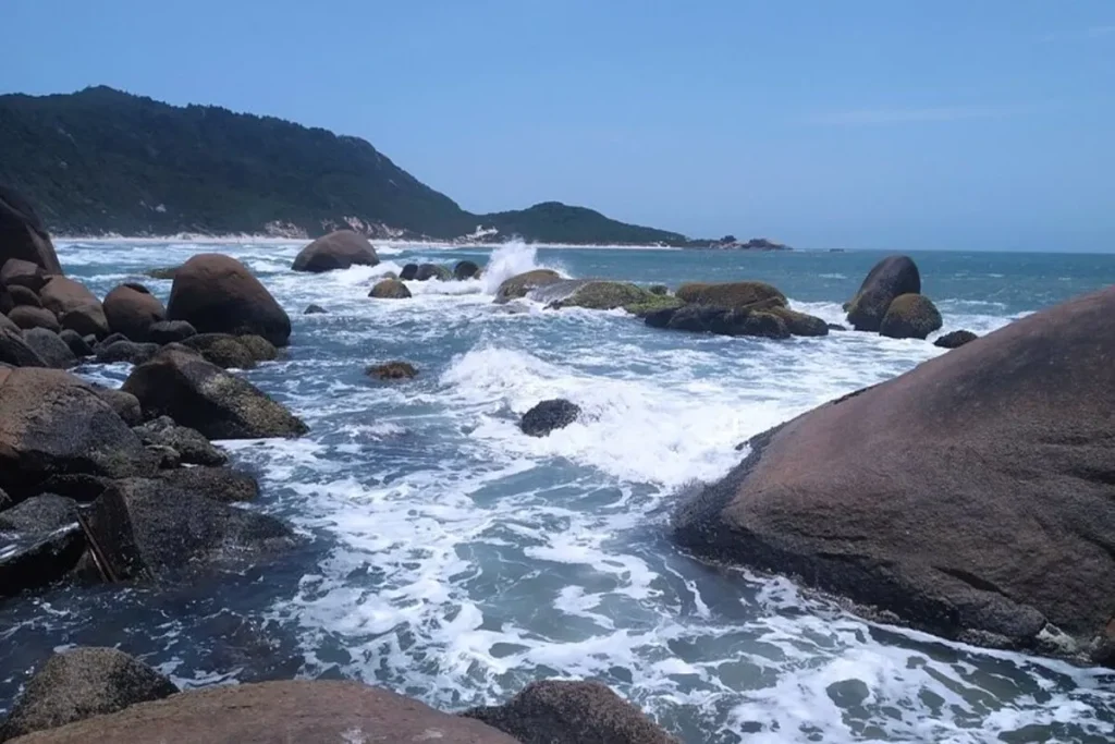 Mar agitado com ondas quebrando em grandes pedras e rochas em uma praia isolada de Florianópolis, com montanhas de mata atlântica ao fundo sob um céu azul.