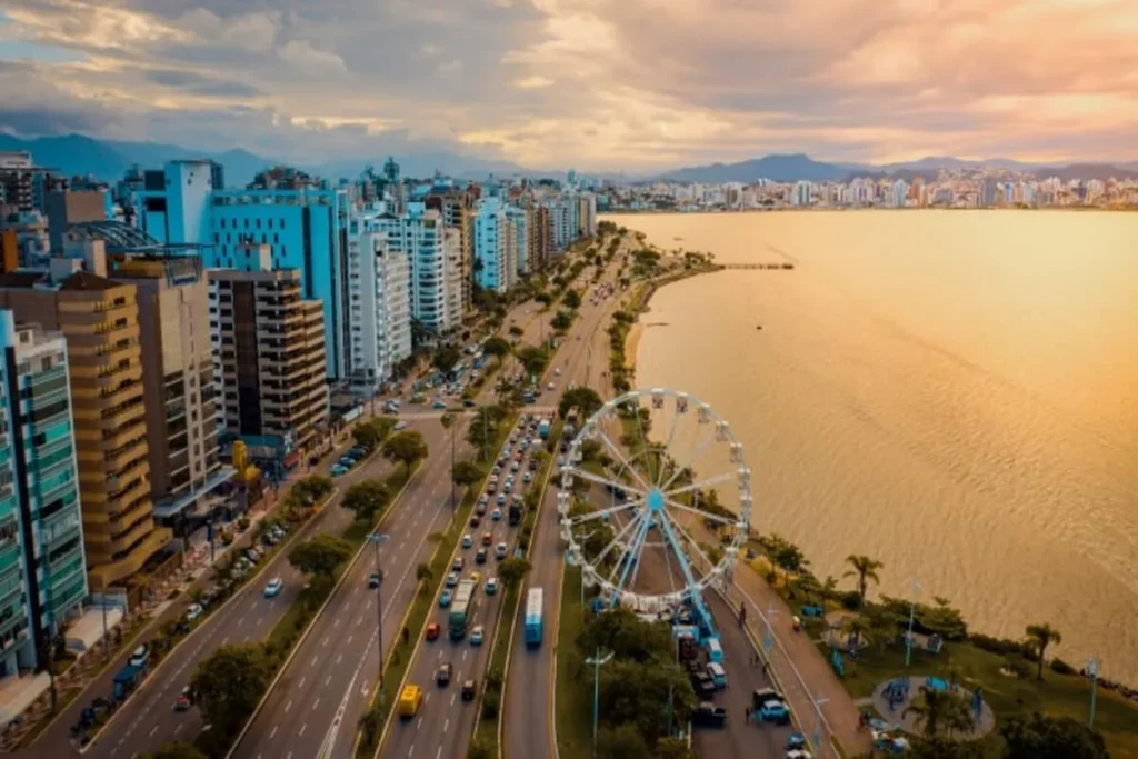 Vista aérea de Florianópolis com a orla iluminada ao entardecer, prédios modernos, roda-gigante e a Avenida Beira-Mar Norte, próxima à prefeitura de Florianópolis.
