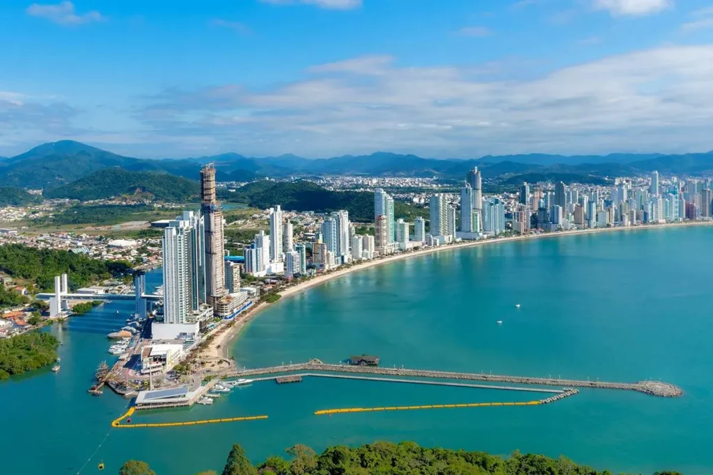 Vista aérea panorâmica da orla urbana de Florianópolis, com uma densa fileira de arranha-céus modernos (prédios brancos e espelhados) alinhados na costa de uma baía de água turquesa. Em primeiro plano, um cais/trapiche se estende sobre a água, com embarcações e um píer. Montanhas verdes formam a paisagem de fundo. A imagem ilustra o desenvolvimento urbano de Florianópolis, uma responsabilidade da Prefeitura.