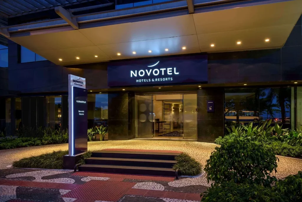 Entrada iluminada do Hotel Novotel em Florianópolis à noite, com fachada moderna, letreiro luminoso e jardins bem cuidados.