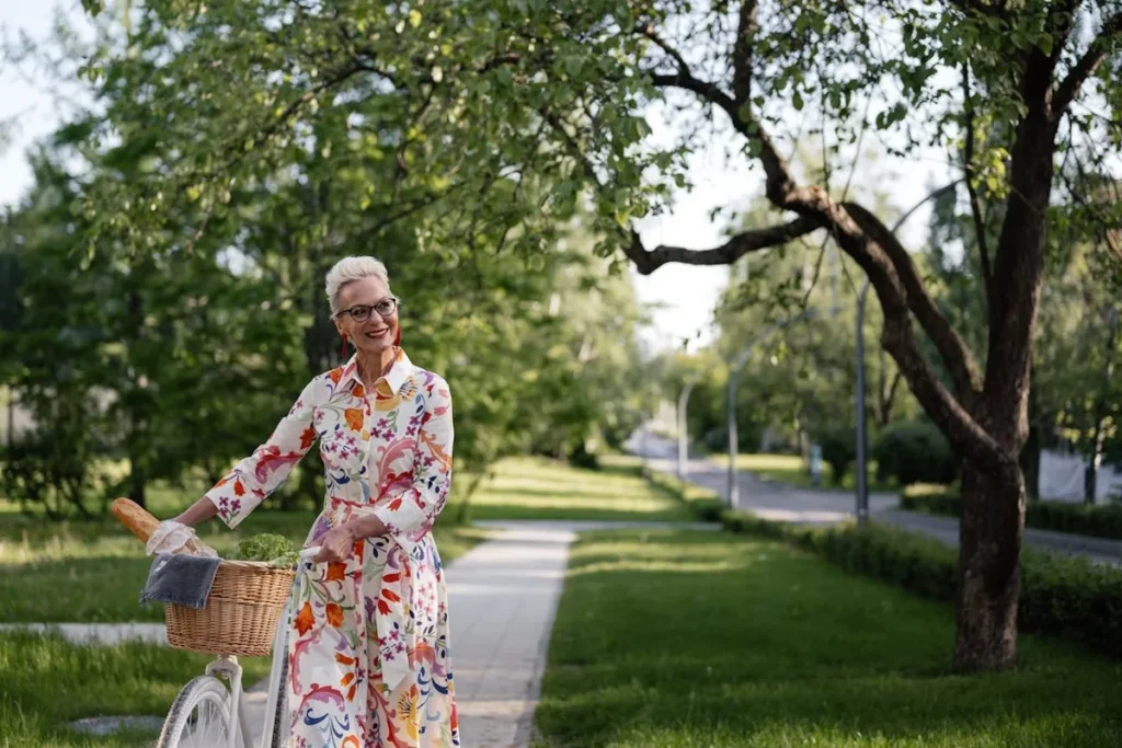 Mulher idosa elegante com cabelo branco e óculos, vestindo um vestido floral, em uma ciclovia com uma bicicleta e cesta cheia.