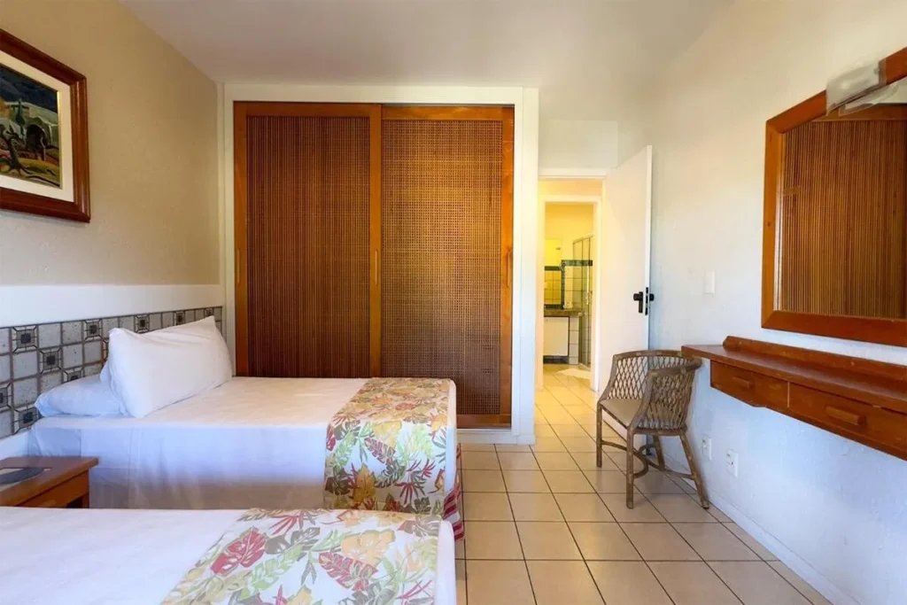 Quarto de hotel duplo no Costão do Santinho, Florianópolis, com duas camas de solteiro, armário de palha trançada e detalhes em madeira. A decoração é simples, com motivos florais tropicais nas roupas de cama.