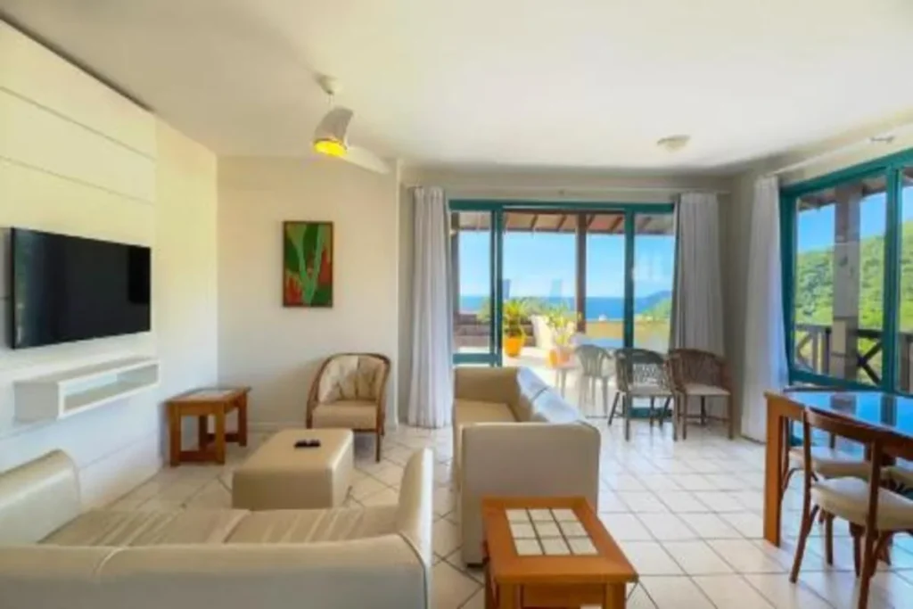 Sala de estar de uma suíte de resort no Costão do Santinho, Florianópolis, com decoração clara, sofá bege e grandes janelas voltadas para uma varanda com vista para o mar