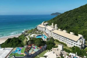 Vista aérea de drone do Costão do Santinho Resort, em Florianópolis. O complexo hoteleiro fica aninhado na colina, com piscinas, área de lazer infantil e acesso direto à praia e ao mar.