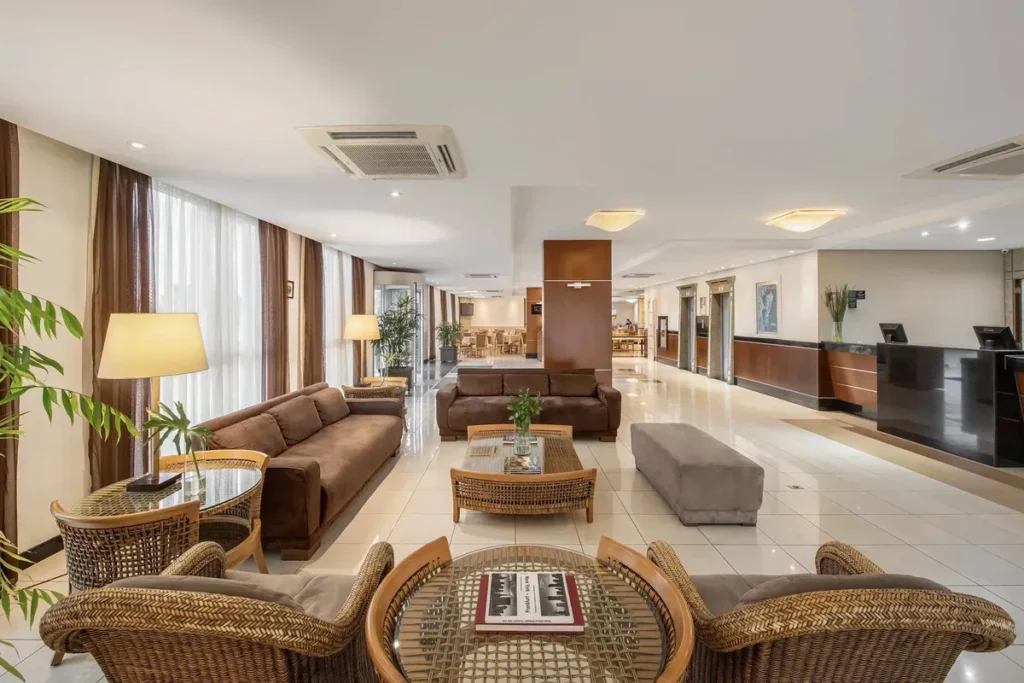 O espaçoso e bem iluminado lobby e lounge do Hotel Intercity Florianópolis, apresentando uma decoração moderna com sofás de suede marrom, poltronas de vime, mesas de centro de vidro e piso de porcelanato branco. A imagem mostra a área de estar com plantas decorativas, a recepção ao fundo e transmite uma atmosfera profissional e acolhedora na entrada do hotel.