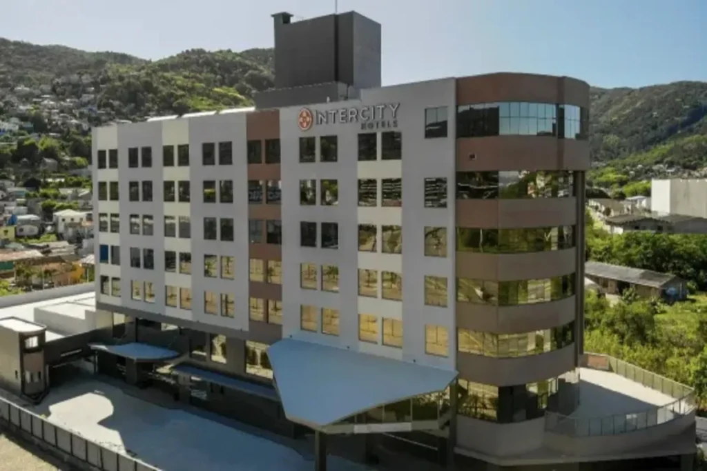Um interior de quarto moderno, associado ao Hotel Intercity Florianópolis, apresentando uma cama de casal com cabeceira em parede texturizada azul-acinzentada, piso de madeira e uma banheiro visível através de uma divisória de vidro. O quarto inclui uma TV de tela plana montada na parede, um pendente de design e cortinas escuras para maior privacidade, destacando o design contemporâneo e funcional.