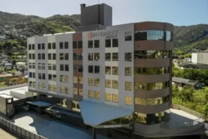 Um interior de quarto moderno, associado ao Hotel Intercity Florianópolis, apresentando uma cama de casal com cabeceira em parede texturizada azul-acinzentada, piso de madeira e uma banheiro visível através de uma divisória de vidro. O quarto inclui uma TV de tela plana montada na parede, um pendente de design e cortinas escuras para maior privacidade, destacando o design contemporâneo e funcional.