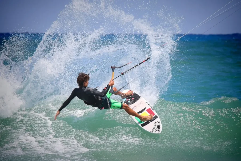 Jovem atleta de kitesurf com roupa preta e verde, saltando radicalmente em uma onda, criando um grande splash de água no mar.