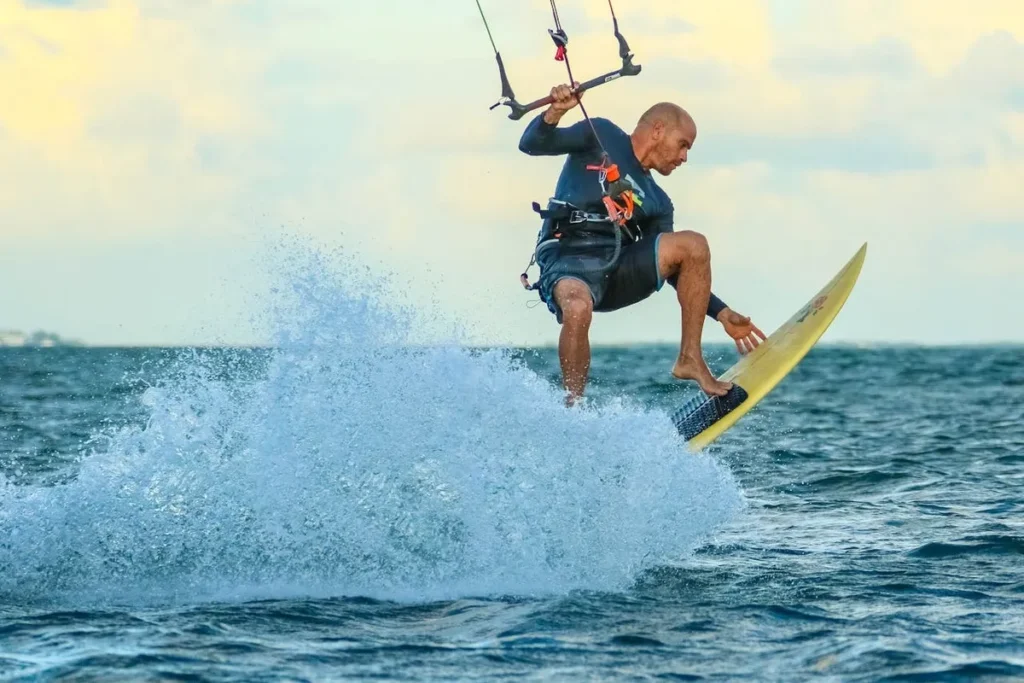 Homem calvo fazendo kitesurf, saltando sobre a água do mar com uma prancha amarela e segurando a barra da pipa.