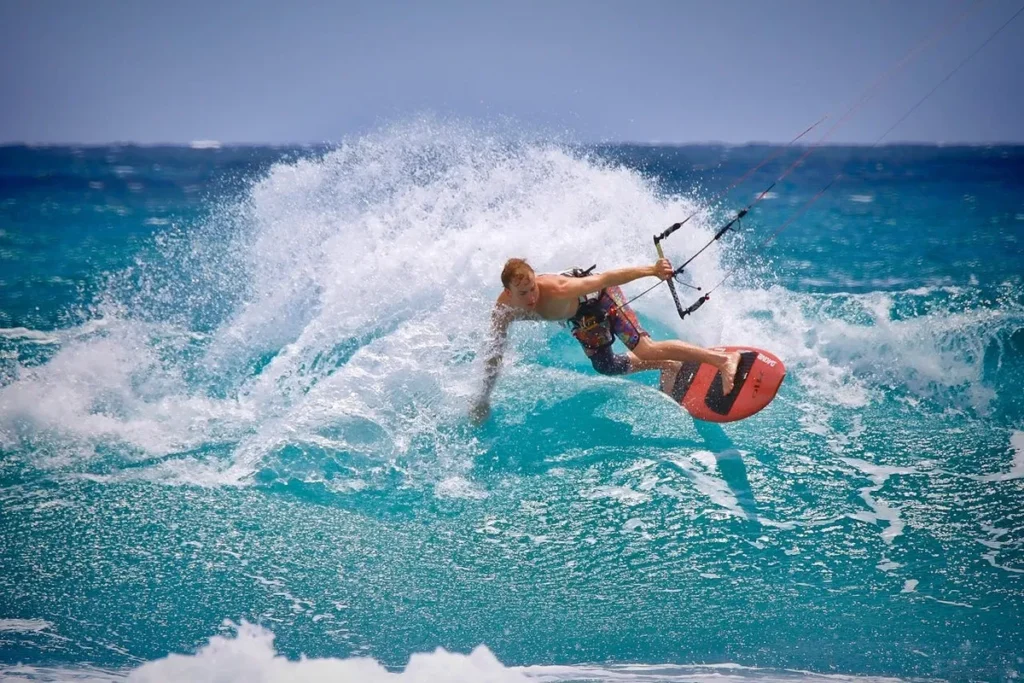 Jovem atleta de kitesurf fazendo um salto sobre uma onda no mar azul, com prancha vermelha e calção estampado.