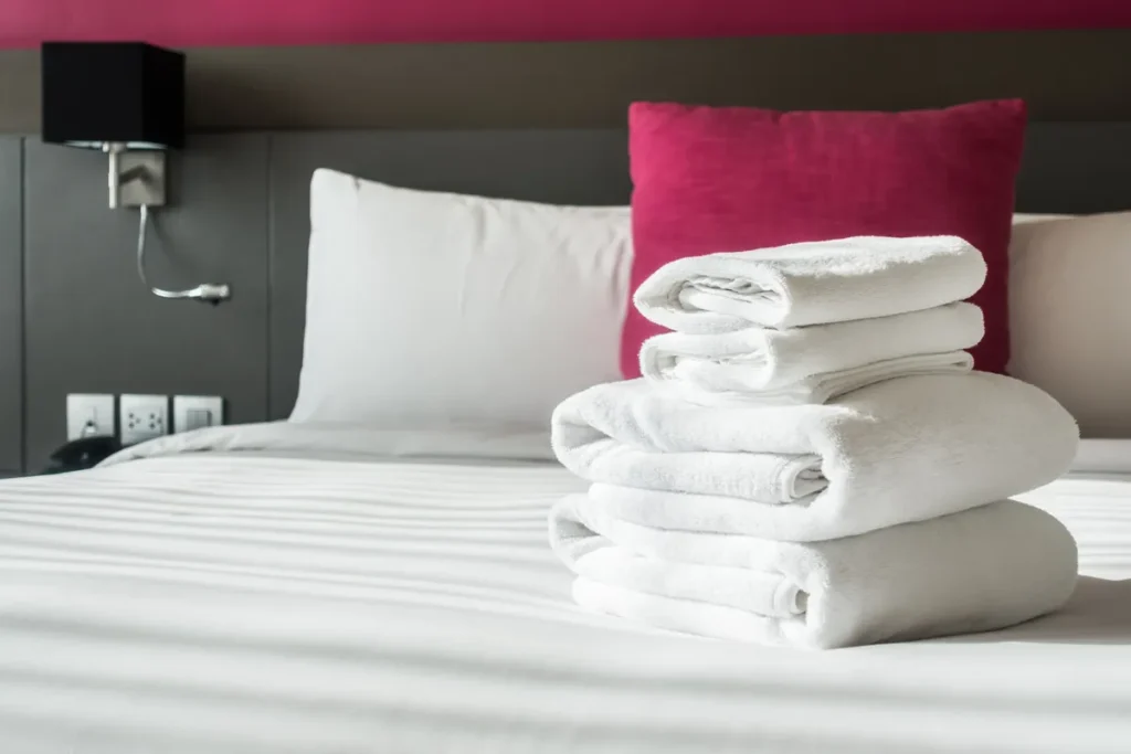Um close-up de uma pilha de toalhas brancas e limpas dobradas sobre uma cama de casal impecável em um quarto de hotel ou motel em Florianópolis. O fundo mostra travesseiros brancos e um grande travesseiro de cor magenta ou rosa choque, com uma cabeceira cinza-escura e um abajur preto. A imagem destaca a qualidade e a higiene das acomodações.