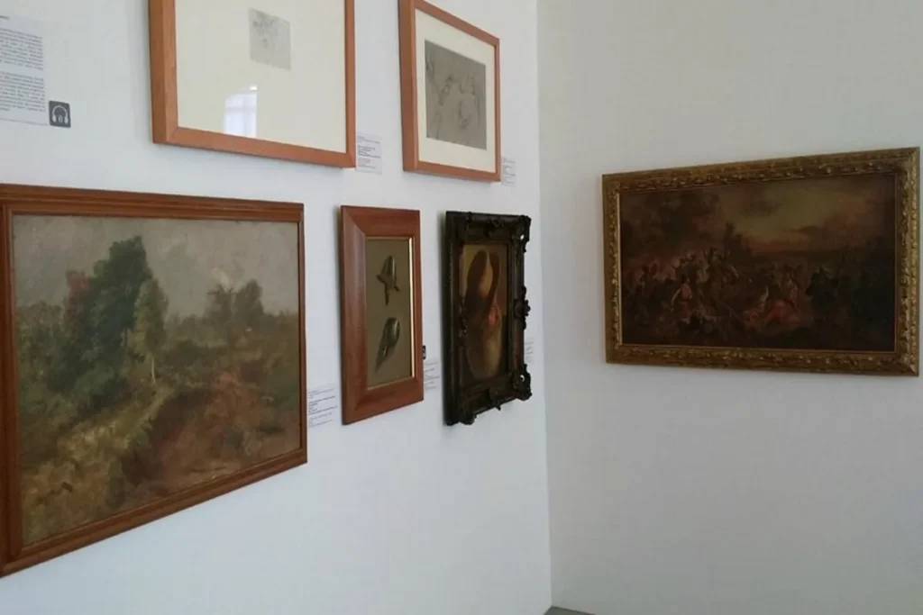Quadros e pinturas emolduradas de diferentes tamanhos e estilos (paisagens e retratos) pendurados em uma parede branca de museu.