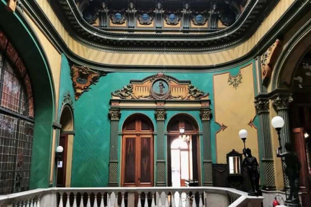 Interior luxuoso de um museu em Florianópolis com teto abobadado, paredes verdes-turquesa e amarelas, sacadas e estátuas escuras.