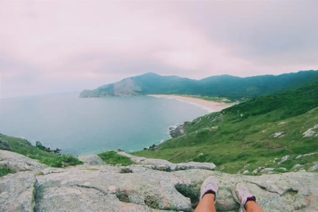 Vista panorâmica de uma praia de areia em curva, com mata verde na colina, e pessoa sentada nas pedras em primeiro plano, na Trilha do Saquinho.