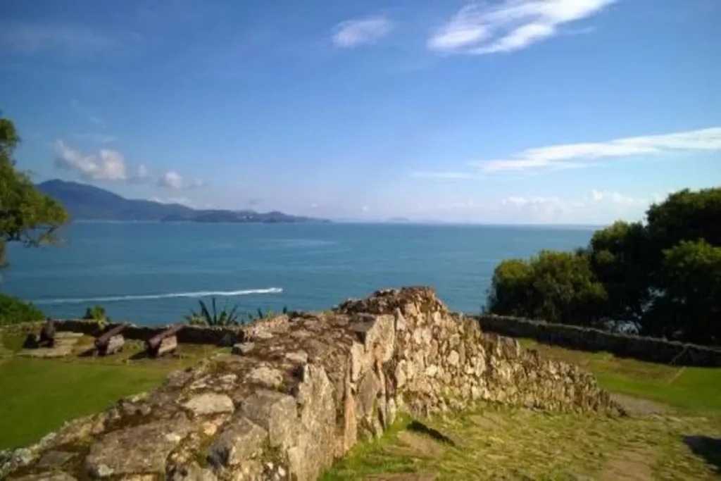 Vista do mar azul e montanhas a partir de uma muralha de pedras em ruínas, com canhões de ferro no gramado, representando uma fortaleza na costa.