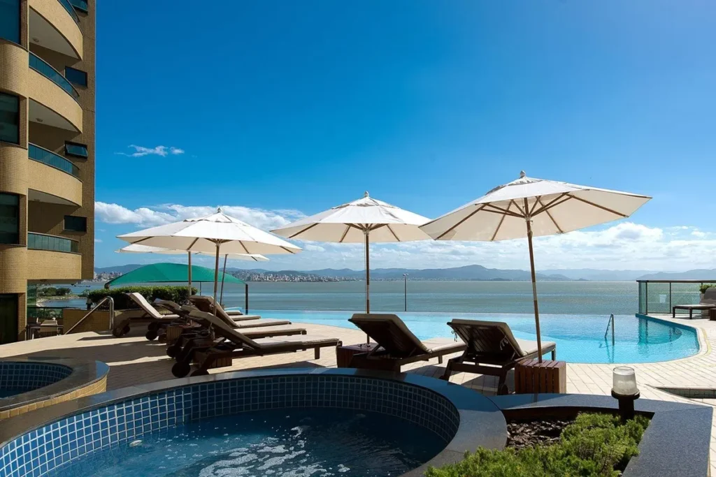 Piscina do Hotel Majestic em Florianópolis com espreguiçadeiras, guarda-sóis brancos e vista panorâmica para o mar e montanhas sob um céu azul.