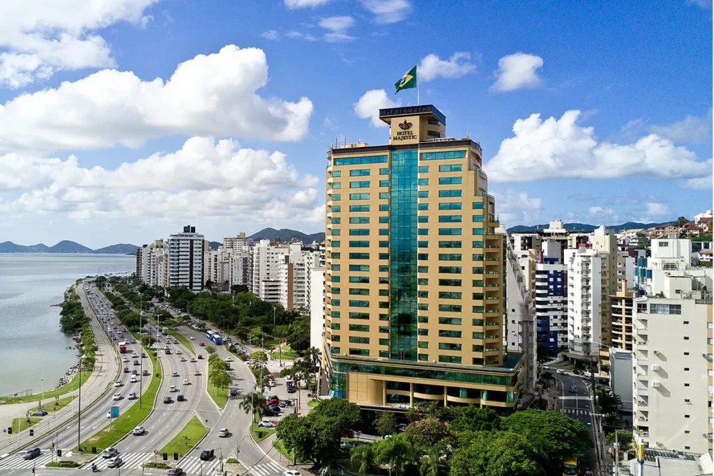 Vista aérea do Hotel Majestic em Florianópolis, com fachada moderna e localização privilegiada na Beira-Mar Norte, cercado por prédios e com o mar ao fundo.