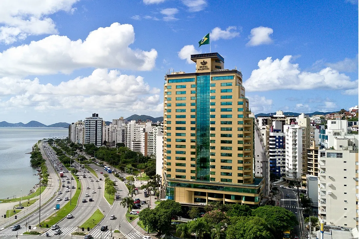 Vista aérea do Hotel Majestic em Florianópolis, com fachada moderna e localização privilegiada na Beira-Mar Norte, cercado por prédios e com o mar ao fundo.