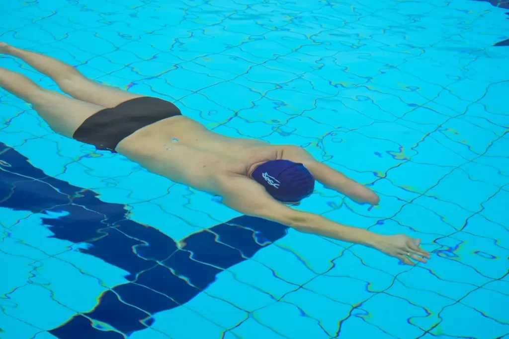 Nadador usando touca azul e sunga preta deslizando de bruços na piscina azul clara, com os braços estendidos à frente.