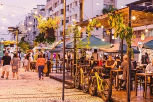 Calçadão em Florianópolis com bares ao ar livre, mesas ocupadas, bicicletas amarelas estacionadas e pessoas caminhando ao entardecer sob luzes decorativas.
