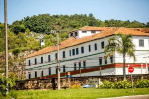 Prédio branco da Penitenciária de Florianópolis com telhado vermelho, cercado por muro de pedra e localizado próximo a área verde e morro com vegetação.